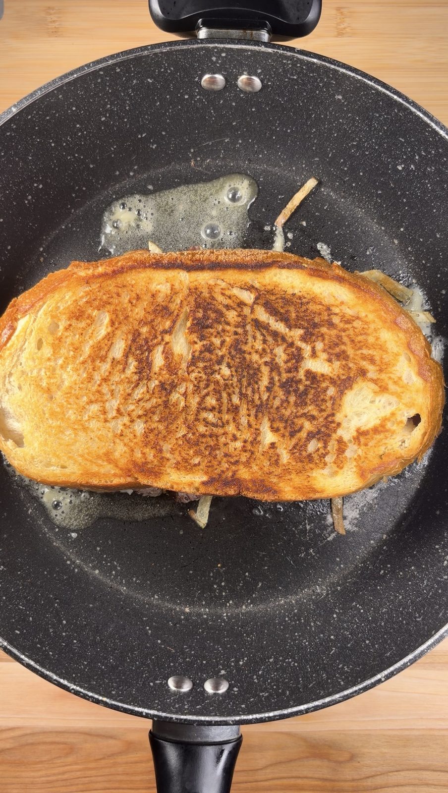 The assembled sandwich toasting in a black pan, top slice golden brown in melted butter