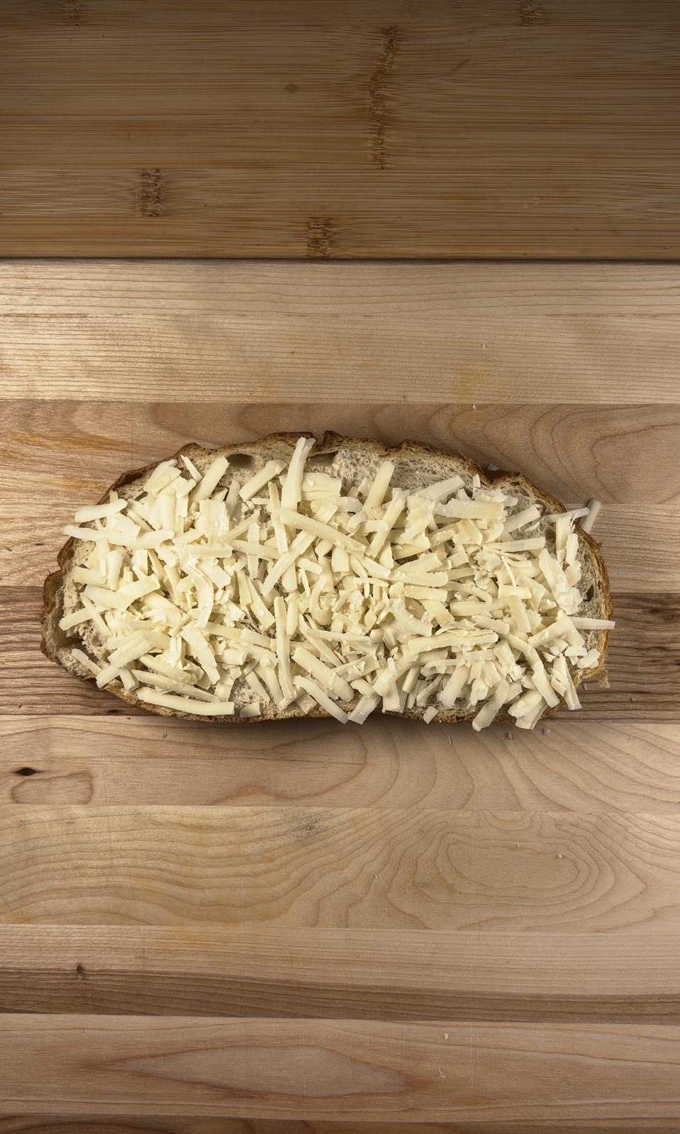 A slice of sourdough covered edge to edge with shredded gruyère on a wooden board
